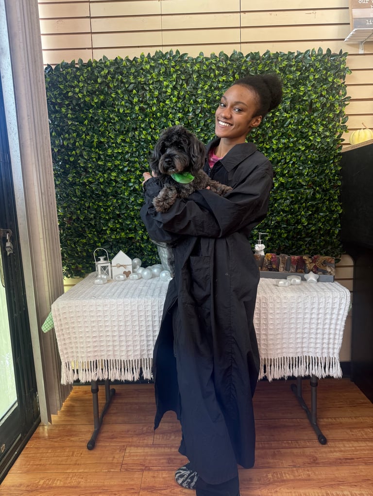 Destiny, professional pet groomer with 5 years experience, holding a black dog in front of a green plant backdrop